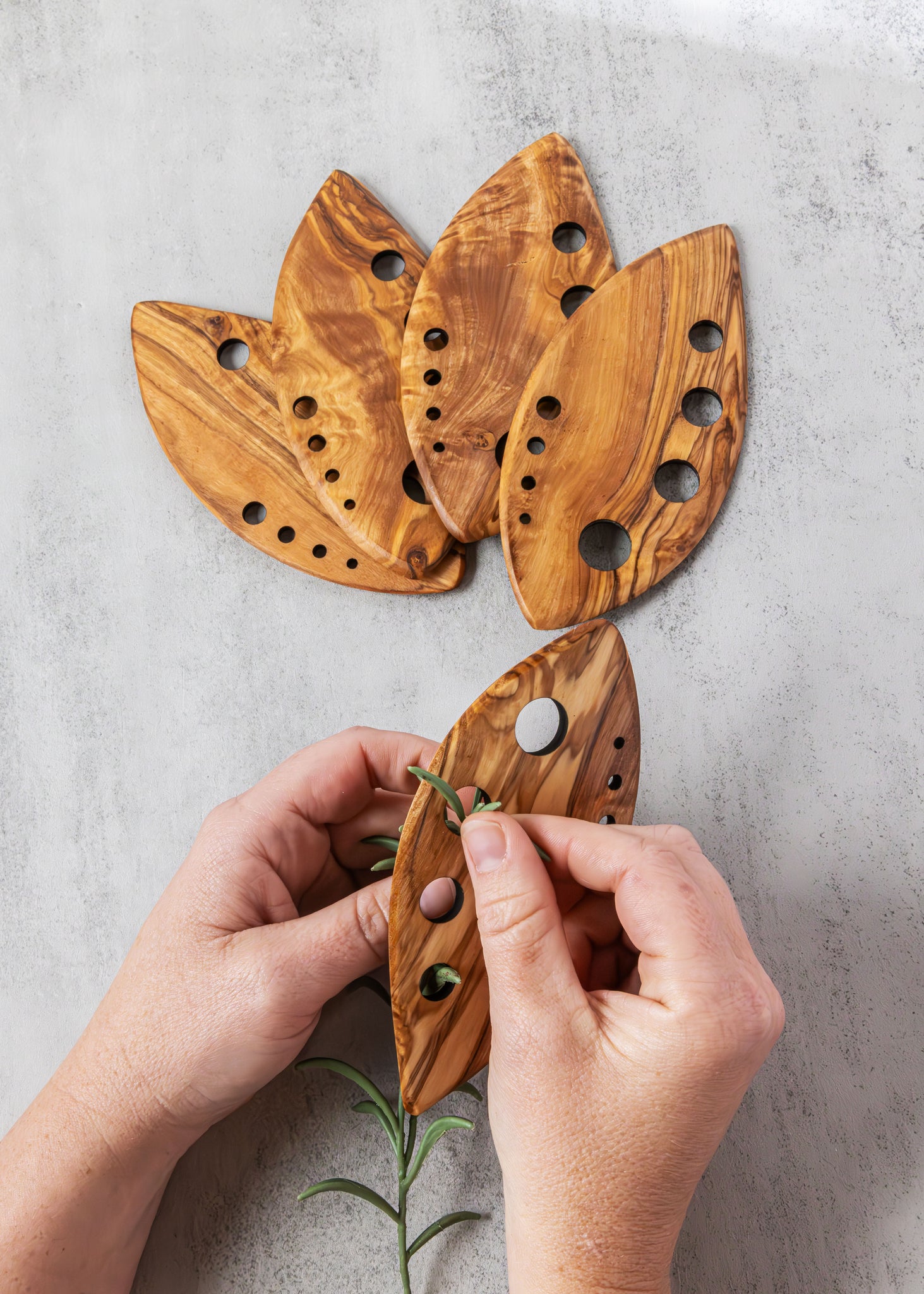 Wooden leaf-shaped plant hanger with hands holding a sprig of rosemary against a light gray background